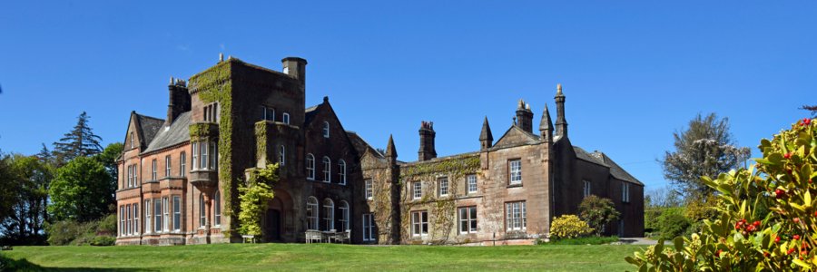 A large red sandstone house with ivy climbing its tower, seen across a mown lawn under a clear sky