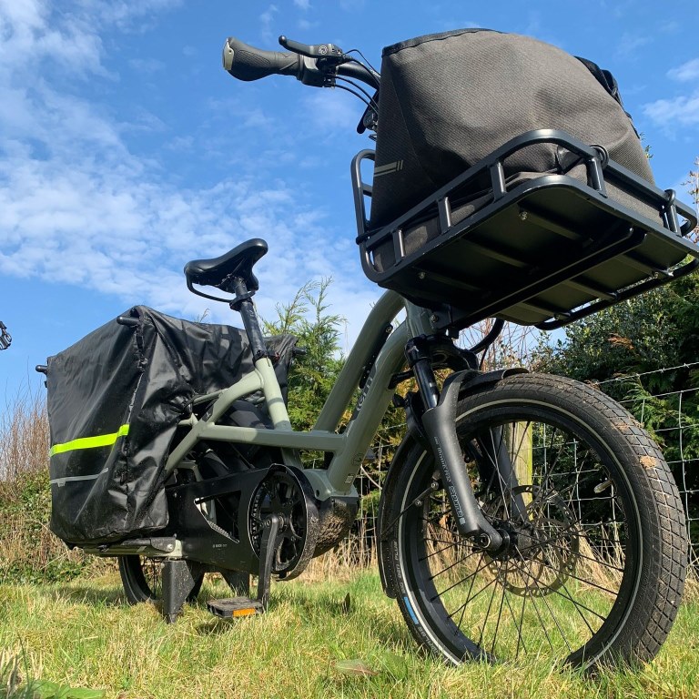 One of ACT’s electric cargo bikes standing on grass beside a wire fence, with a large black pannier and a loaded front rack, under a bright blue sky