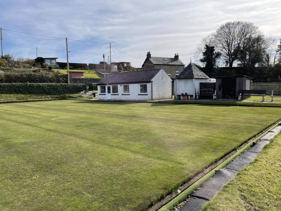 The Auchencairn bowling green in low winter sun, with the white clubhouse and the village houses on the hill behind
