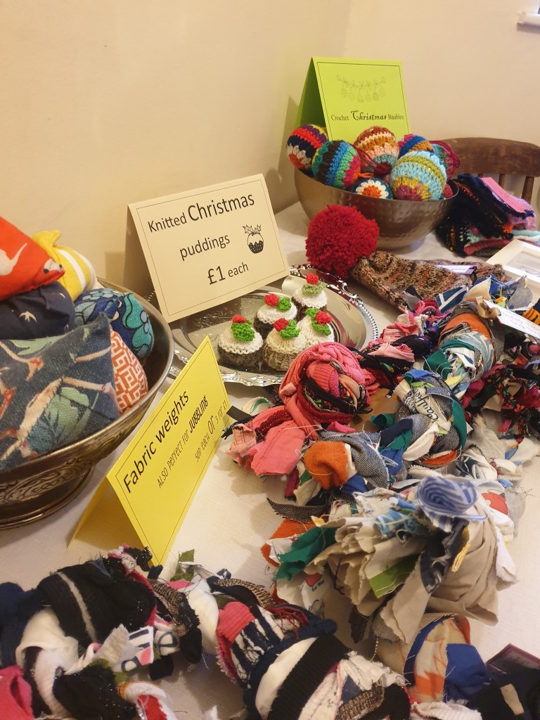 A table of handmade crafts for sale in the hall: knitted Christmas puddings, crocheted baubles in a metal bowl, and fabric weights, with handwritten price cards